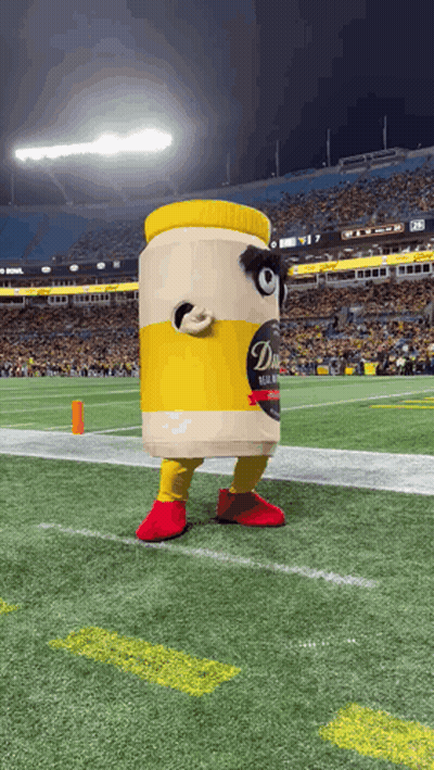 A Mascot Dressed As A Jar Of Mustard Is Standing On A Football Field . GIF