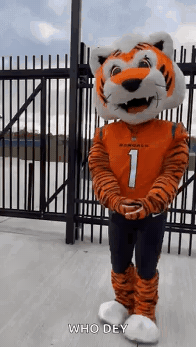 A Mascot Dressed As A Tiger Is Standing In Front Of A Fence With His Arms Crossed . GIF