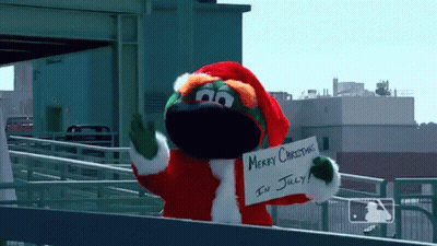 A Mascot Dressed In A Santa Suit Is Holding A Sign That Says Merry Christmas In July GIF