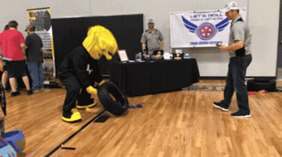 A Mascot Is Rolling A Tire In Front Of A Sign That Says Let 'S Roll GIF