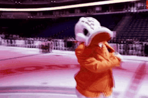 A Mascot Is Standing On A Hockey Rink Wearing An Orange Jacket And A White Hat GIF