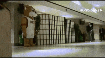 A Mascot Wearing A Bucks Jersey Is Standing In A Hallway .. GIF