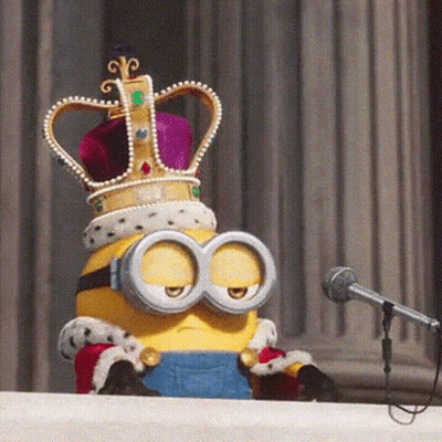 A Minion Wearing A Crown And Goggles Is Sitting At A Podium In Front Of A Microphone GIF