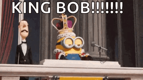 A Minion Wearing A Crown Is Sitting At A Table With A Microphone . GIF