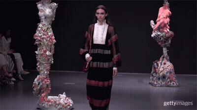 A Model Walks Down The Runway At A Fashion Show In Front Of Sculptures GIF