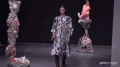 A Model Walks Down The Runway At A Fashion Show In Front Of Statues GIF