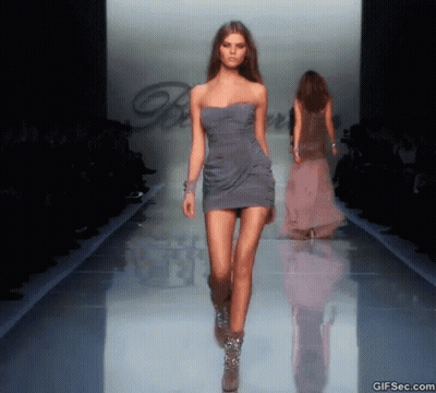 A Model Walks Down The Runway At A Fashion Show Wearing A Grey Dress GIF