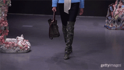A Model Walks Down The Runway At A Fashion Show While Holding A Purse And Wearing Green Boots GIF