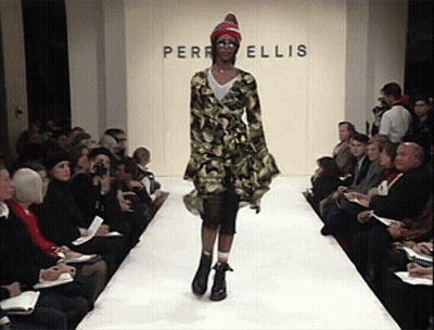 A Model Walks Down The Runway In Front Of A Wall That Says Perri Ellis GIF
