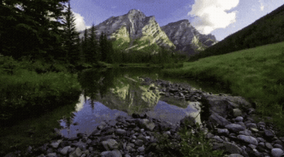 A Mountain Is Reflected In A Lake With Rocks In The Foreground GIF