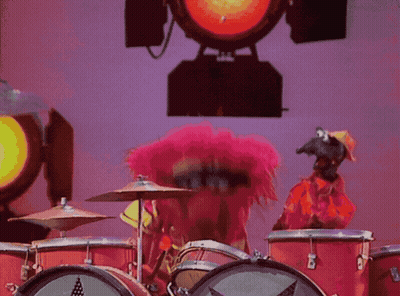 A Muppet With Pink Hair Plays Drums In Front Of A Red Light GIF