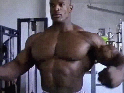 A Muscular Man Is Standing In A Gym With His Arms Outstretched And Looking At The Camera . GIF