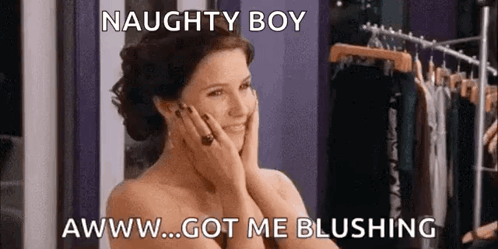 A Naked Woman Is Standing In Front Of A Clothes Rack With Her Hands On Her Face And Smiling . GIF