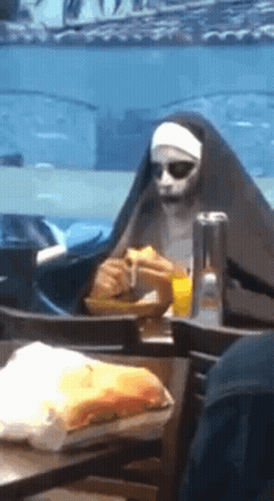 A Nun Is Sitting At A Table Eating A Sandwich And Drinking Orange Juice . GIF