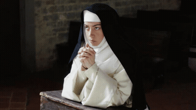 A Nun Sits On A Wooden Table With Her Hands Folded GIF