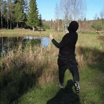 A Person In A Black Hoodie Is Standing In A Field Near A Lake GIF