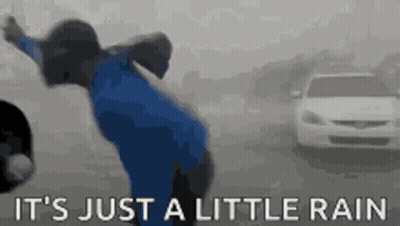 A Person In A Blue Jacket Is Standing In The Rain With A Car In The Background . GIF
