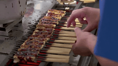 A Person In A Blue Shirt Is Cooking A Row Of Skewers On A Grill GIF