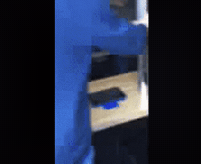 A Person In A Blue Suit Is Standing Next To A Wooden Table With A Cell Phone On It . GIF