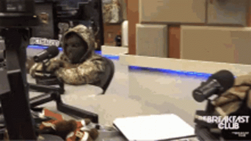 A Person In A Hooded Jacket Is Sitting In Front Of A Breakfast Club Microphone GIF