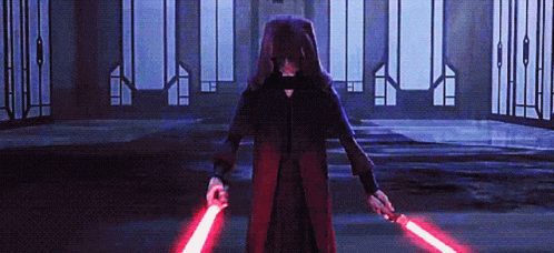 A Person In A Hooded Robe Is Holding Two Red Lightsabers In A Dark Room GIF