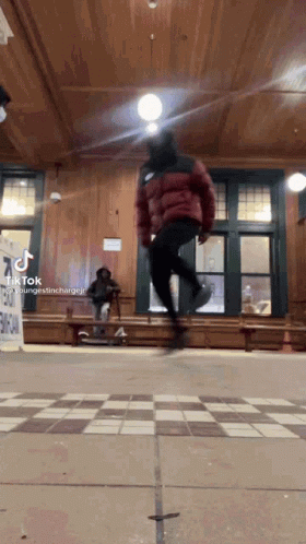 A Person In A Red Jacket Is Jumping In The Air In A Room With A Checkered Floor . GIF