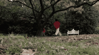A Person In A Red Shirt Is Running In A Park Near A White Bench GIF