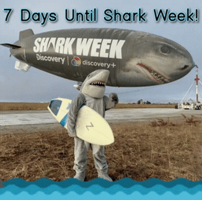 A Person In A Shark Costume Holding A Surfboard In Front Of A Blimp That Says Shark Week GIF