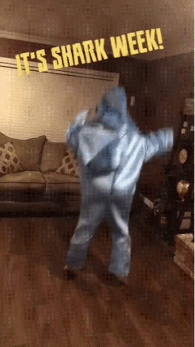 A Person In A Shark Costume Is Dancing In A Living Room With A Sign That Says It 'S Shark Week GIF