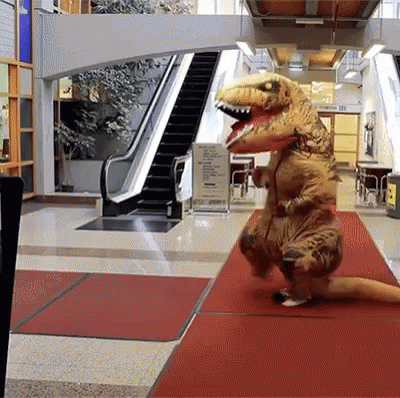 A Person In A T-Rex Costume Is Walking Down A Red Carpet In Front Of An Escalator GIF