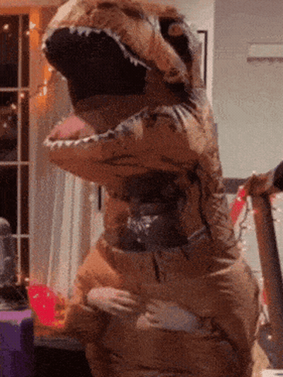 A Person In An Inflatable Dinosaur Costume Is Standing In A Room GIF