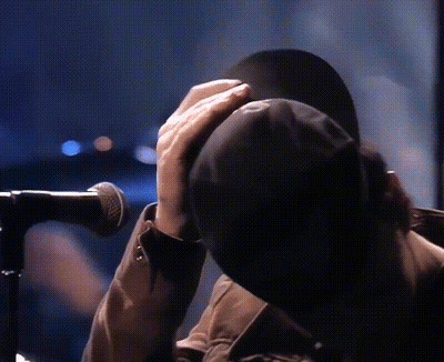 A Person Is Covering Their Face With A Hat While Standing In Front Of A Microphone GIF