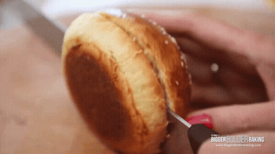 A Person Is Cutting A Hamburger Bun With A Knife And It Says Bigger Bolder Baking On The Bottom Right GIF