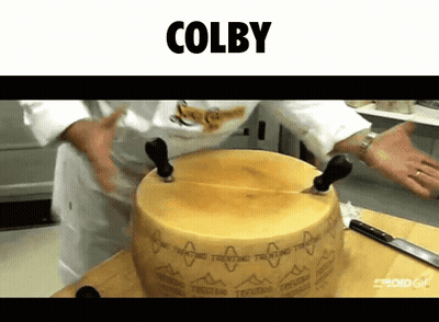 A Person Is Cutting A Large Piece Of Cheese With The Name Colby Written Above Them GIF