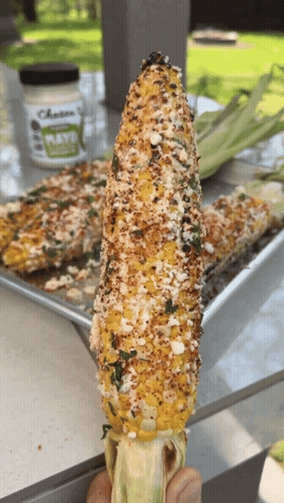 A Person Is Holding A Corn On The Cob With A Jar Of Cheese Sauce In The Background GIF