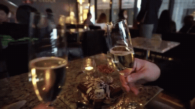 A Person Is Holding A Glass Of Champagne In Front Of A Dessert GIF