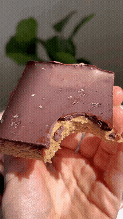 A Person Is Holding A Piece Of Chocolate Covered Dessert With A Bite Taken Out Of It GIF