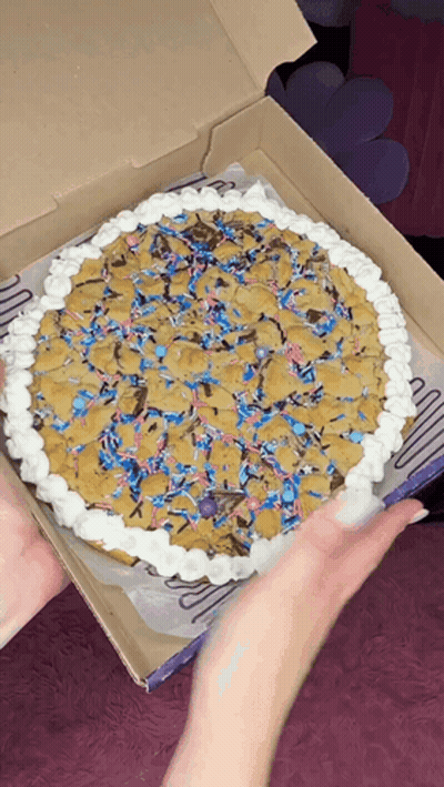 A Person Is Holding A Pizza In A Box That Looks Like A Cookie Cake GIF