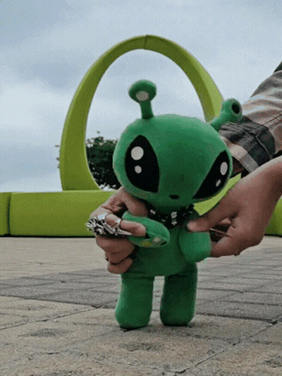 A Person Is Holding A Stuffed Green Alien In Front Of A Green Arch GIF