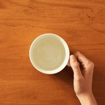 A Person Is Holding A White Cup Of Water On A Wooden Table GIF