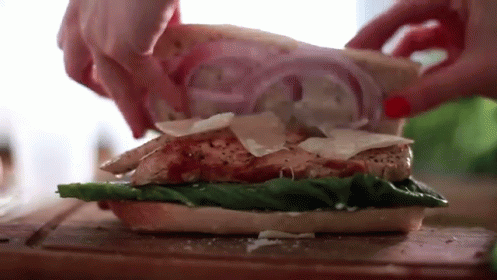 A Person Is Making A Sandwich With Chicken , Lettuce , Cheese And Onions On A Cutting Board . GIF