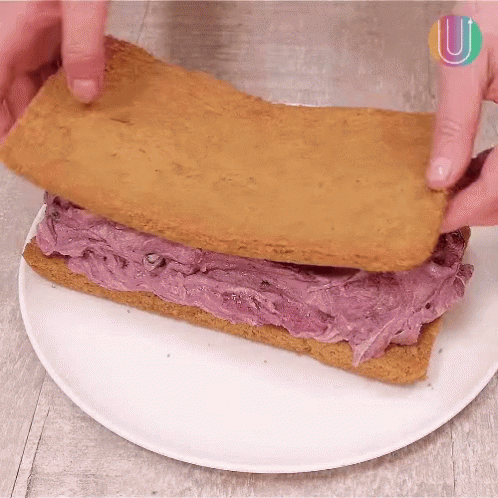 A Person Is Making A Sandwich With Purple Frosting On A White Plate .. GIF
