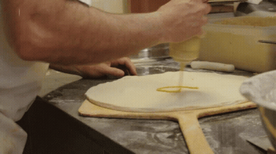 A Person Is Pouring Sauce On A Pizza Dough On A Wooden Paddle GIF