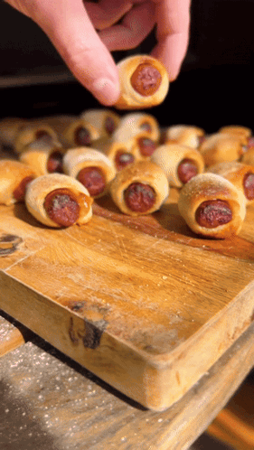 A Person Is Reaching For A Hot Dog Wrapped In A Pastry On A Wooden Cutting Board GIF