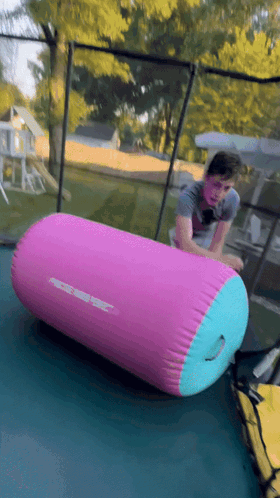 A Person Is Rolling On A Pink And Blue Inflatable Cylinder On A Trampoline GIF