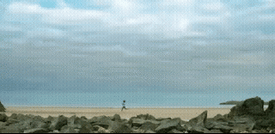 A Person Is Running On A Sandy Beach With Rocks In The Foreground GIF