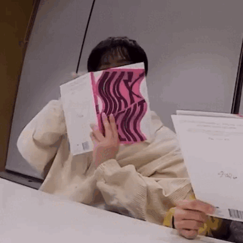 A Person Is Sitting At A Table Covering Their Face With A Pink Book . GIF
