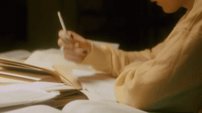 A Person Is Sitting At A Table Writing In A Notebook With A Pencil . GIF