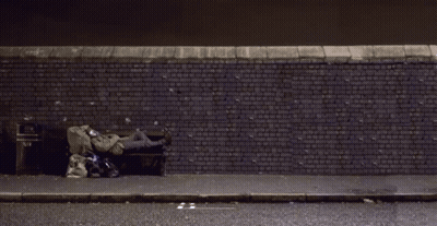 A Person Is Sleeping On A Bench In Front Of A Brick Wall With A Trash Can On The Sidewalk GIF