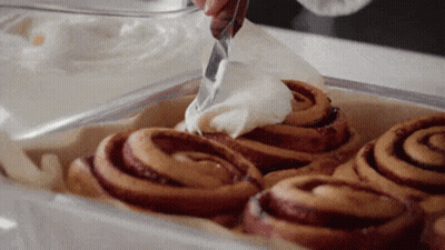 A Person Is Spreading Frosting On Cinnamon Rolls GIF